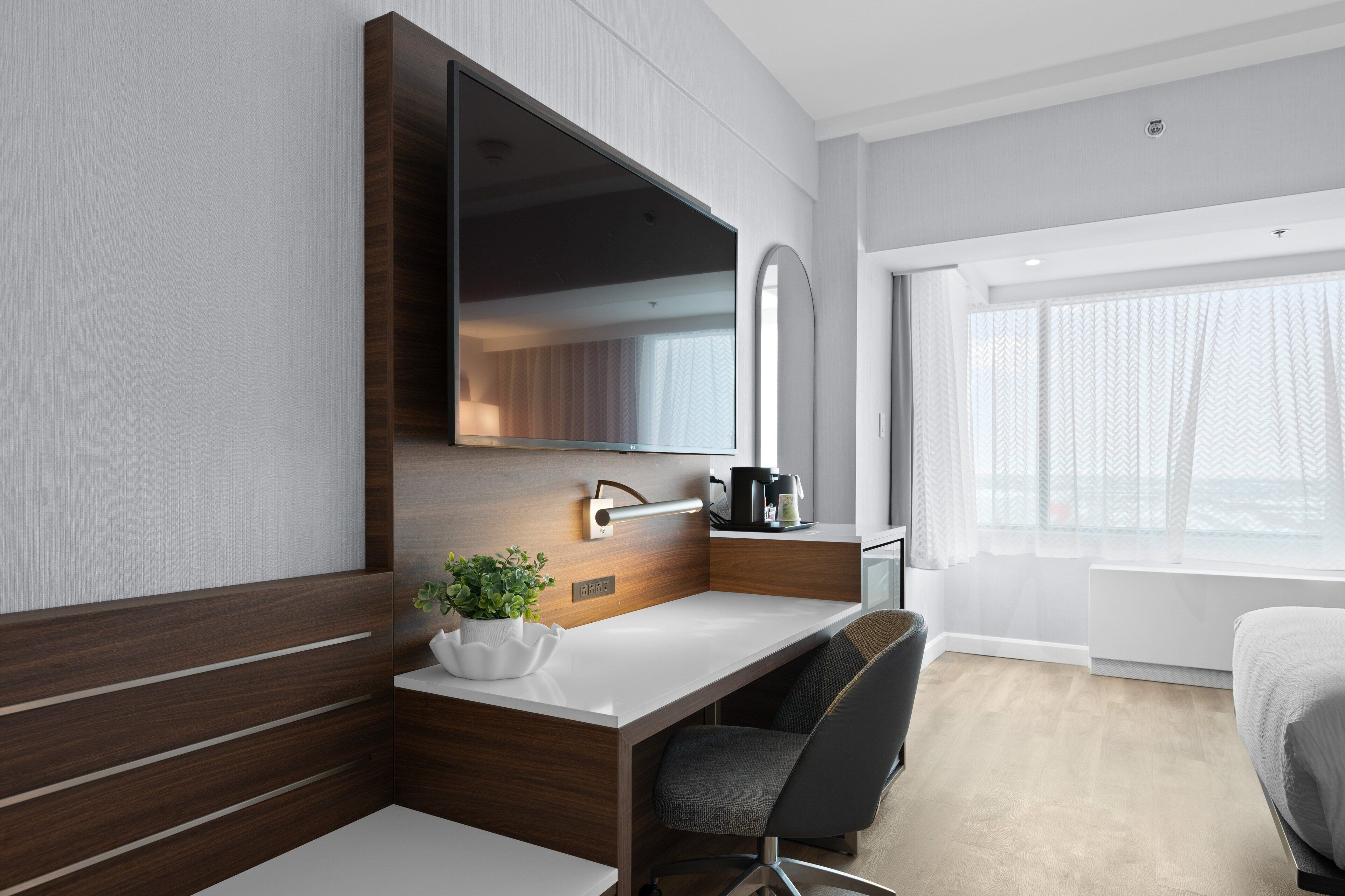 A modern hotel room with a wooden desk, a large mirror, a potted plant, and a bed.