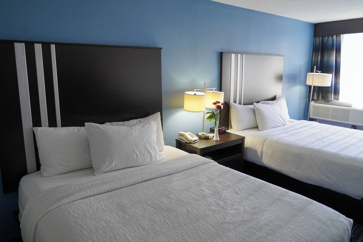 A hotel room with two beds, a nightstand with a lamp, a phone, and a vase with flowers.