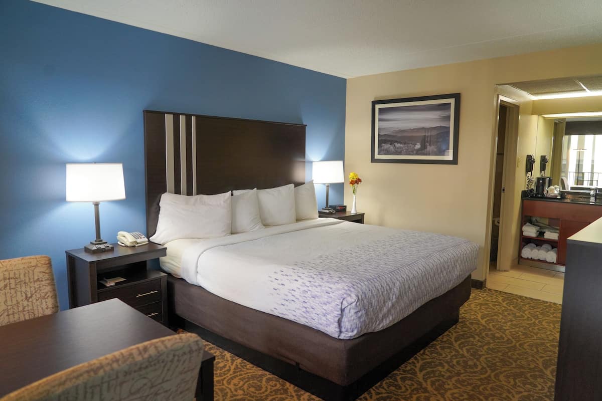 A hotel room with a large bed, two bedside tables with lamps, a chair, a desk, and a bathroom visible through an open door.