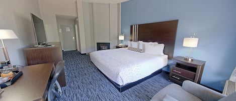 A hotel room with a large bed, a desk with a chair, a bedside table with a lamp, and a wardrobe.