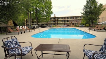 A swimming pool area with lounge chairs and a table, surrounded by trees and a building in the background.