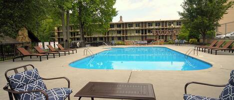A swimming pool area with lounge chairs and a table, surrounded by trees and a building in the background.