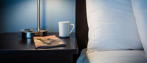 A neatly made bed with white pillows and a white mug on a nightstand. A magazine is also on the nightstand.
