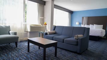 Ein Hotelzimmer mit blauem Teppich, einer Couch, einem Sessel, einem kleinen Tisch und einem Bett mit weißer Bettwäsche.