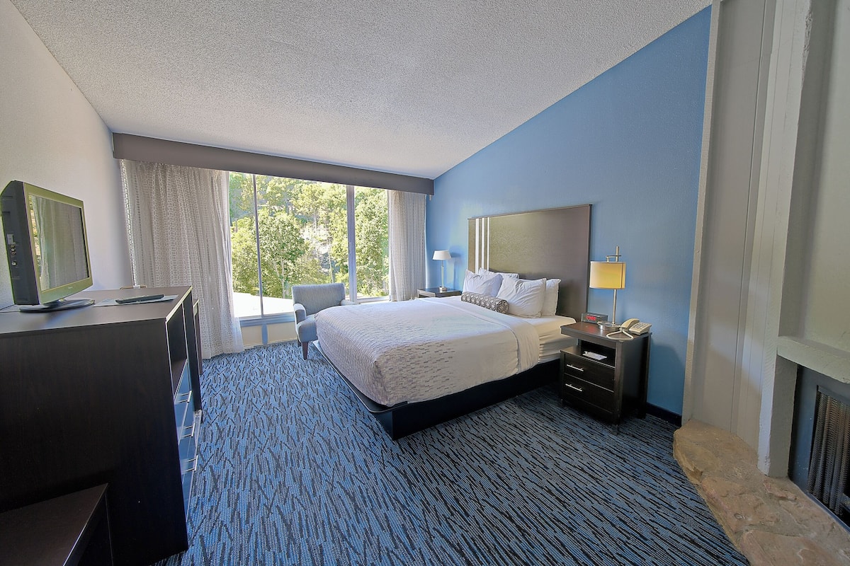 A hotel room with a large bed, a desk with a computer, a chair, a nightstand with a lamp, and a fireplace.