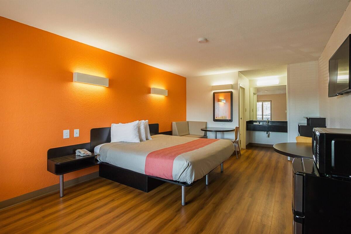 A hotel room with a bed, bedside tables, a desk, a chair, a TV, and a bathroom visible through an open door.
