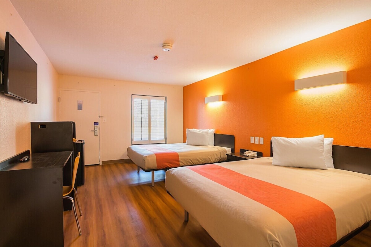 A hotel room with two beds, a desk, a chair, a TV, and a window with blinds.
