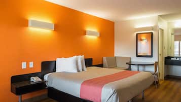 Habitació d'hotel amb una paret taronja, un llit amb llençols beix i vermells, una taula petita amb una cadira, i un mirall sobre la taula.