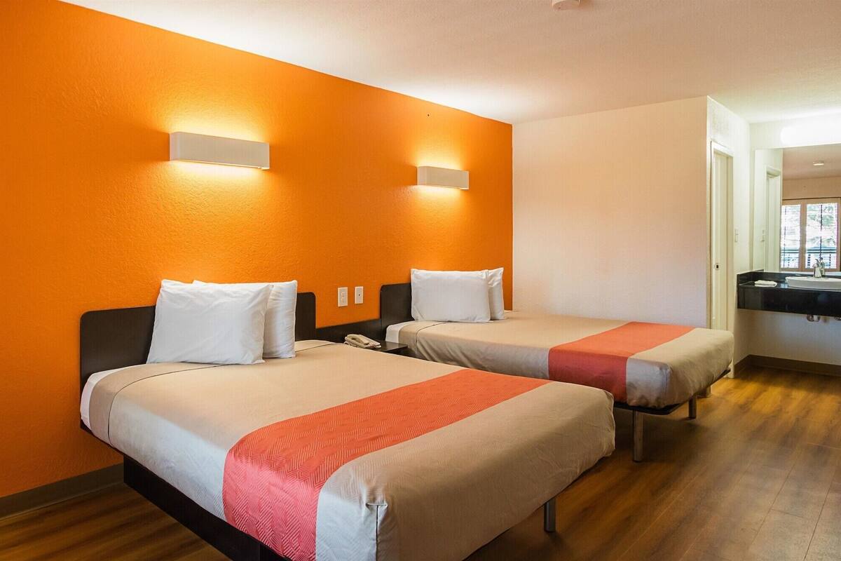 A hotel room with two beds, each with white bedding and a red accent, orange walls, and a small bedside table with a phone.