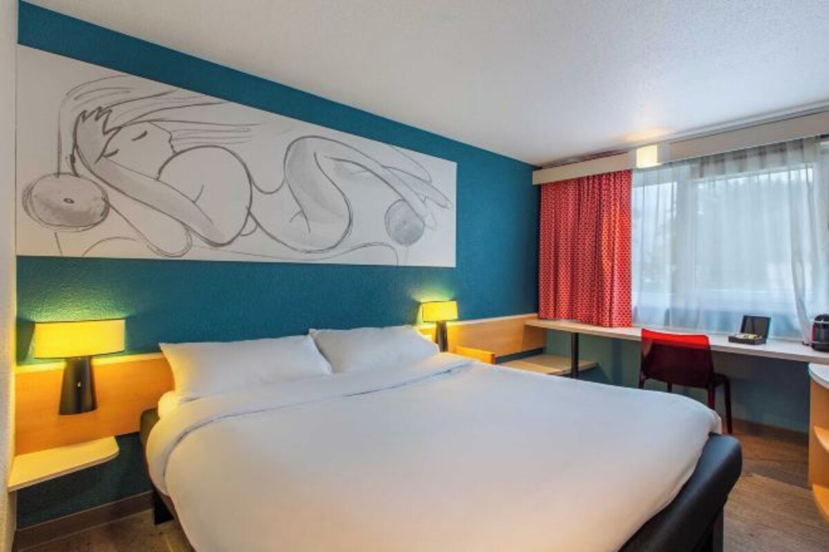 A hotel room with a bed, two bedside lamps, a desk with a chair, a window with curtains, and a wall art piece.