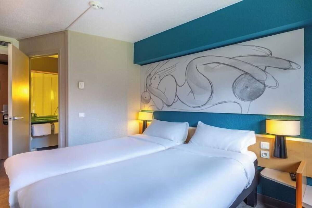 A hotel room with a bed, two bedside lamps, a wall art piece, and a bathroom visible through an open door.