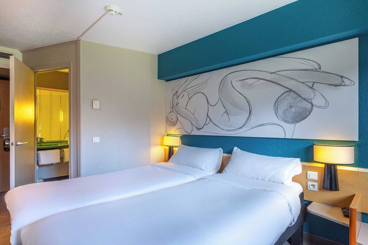A hotel room with a large bed, two bedside lamps, a wall art piece, and a bathroom visible through an open door.