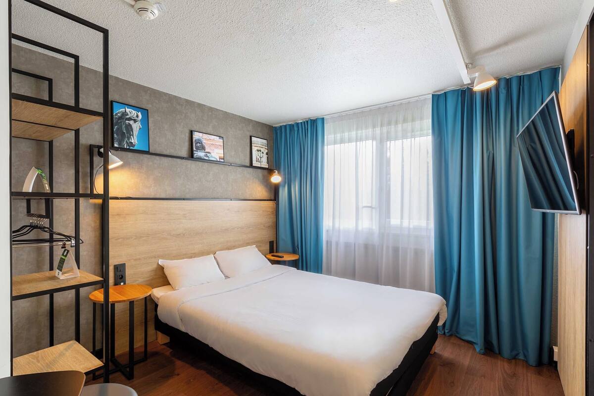 A modern hotel room with a large bed, a wooden headboard, a TV, and a desk with a lamp.