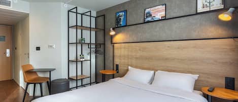 A modern hotel room with a large bed, a desk, a chair, a small table, and a bookshelf.
