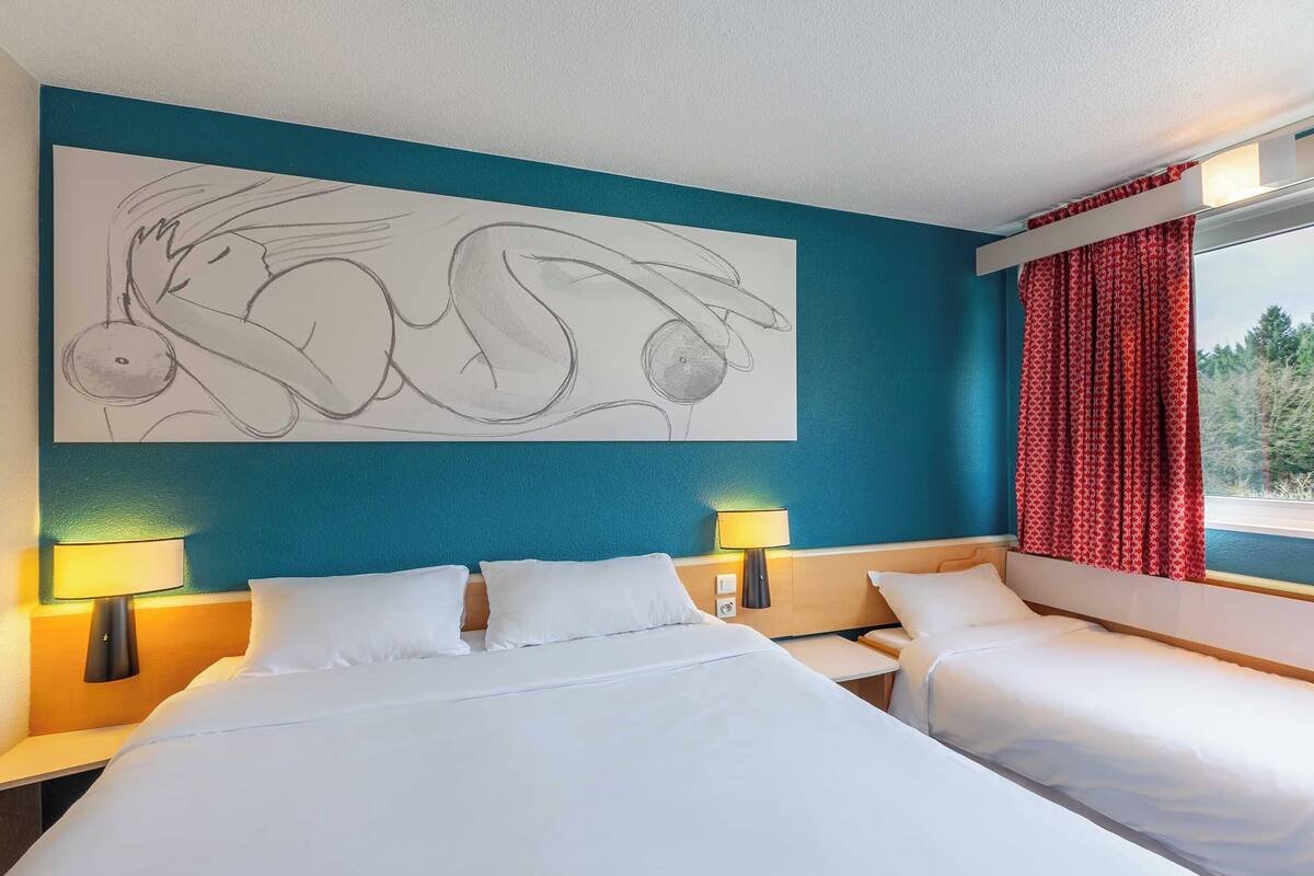 A hotel room with a large bed, two bedside tables, a wall-mounted lamp, and a window with red curtains.