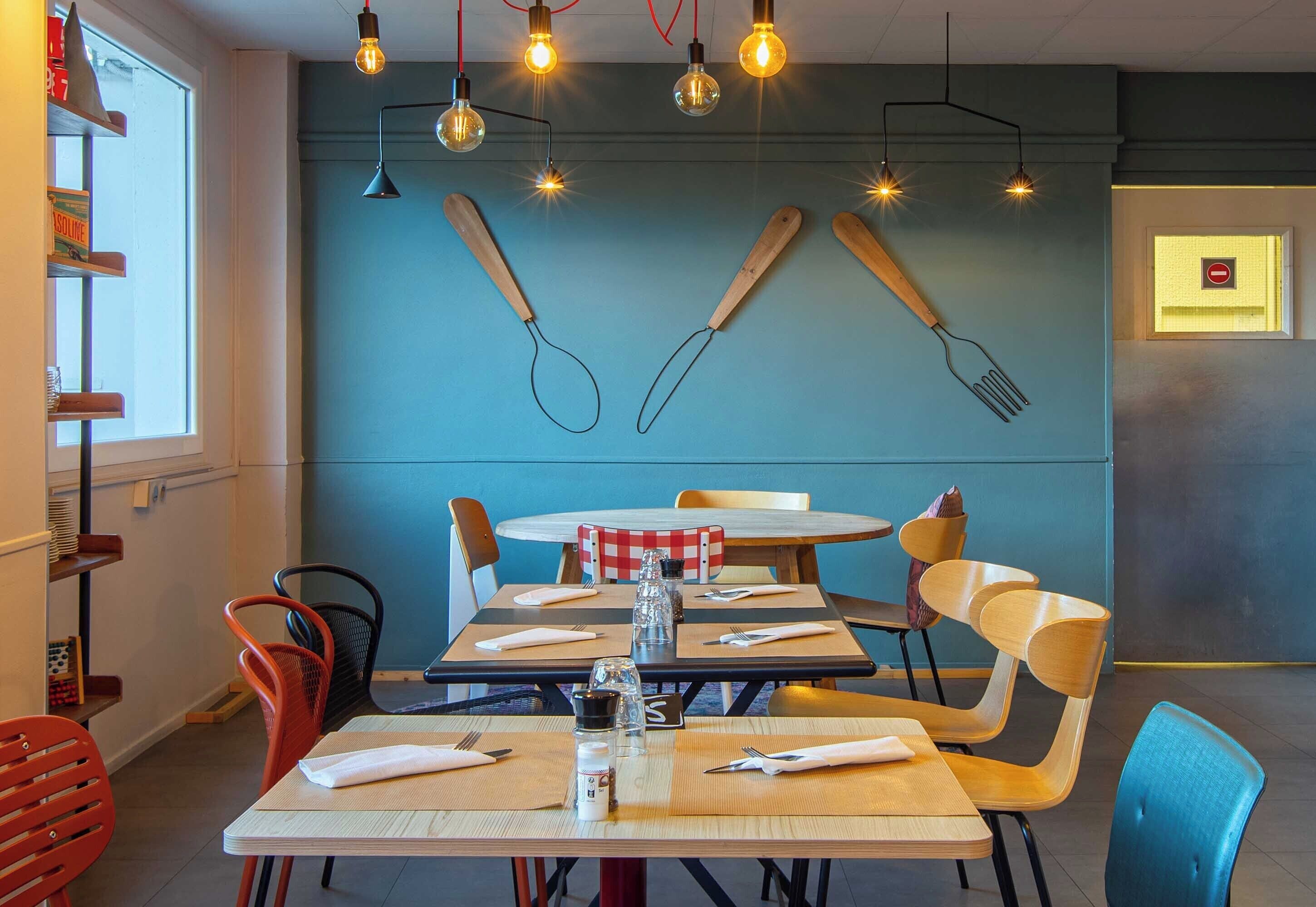 A dining area with wooden tables, chairs, and a blue wall with kitchen utensils as decor.