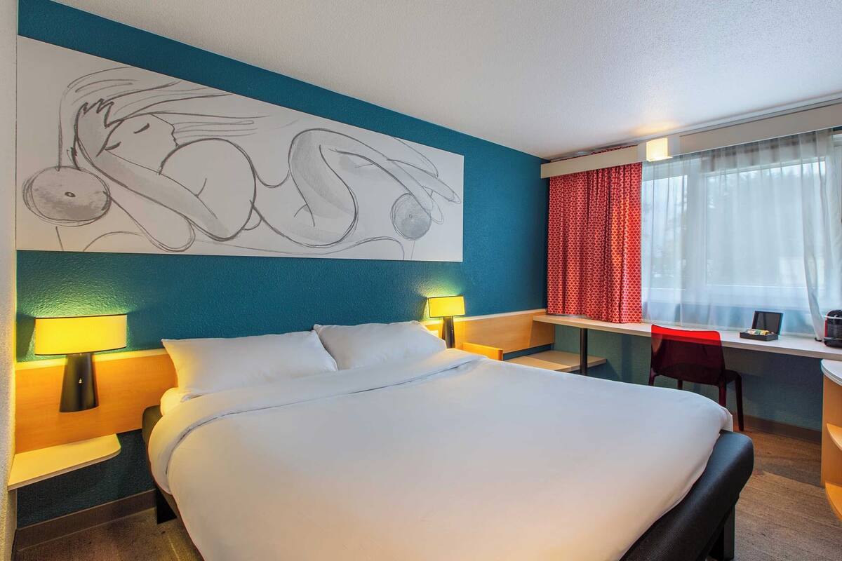 A hotel room with a large bed, two bedside lamps, a desk with a chair, and a window with curtains.