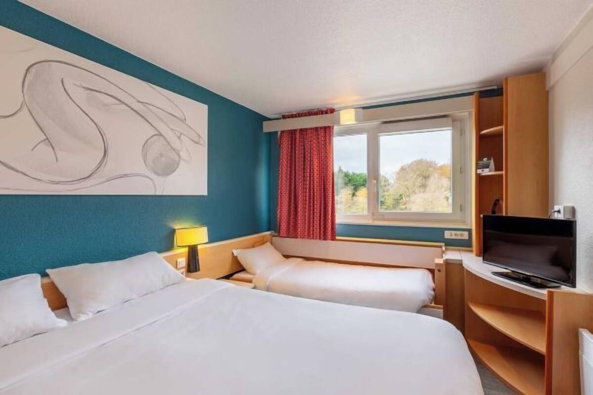 A hotel room with two beds, a TV, a desk, and a large window with curtains.