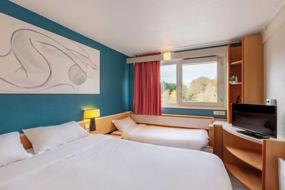 A hotel room with two beds, a TV, a desk, and a large window with curtains.