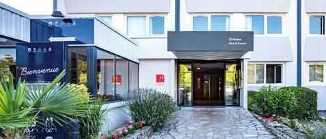 Una entrada de hotel moderna con un camino de piedra que lleva a la puerta, rodeada de plantas decorativas y un cartel de bienvenida.