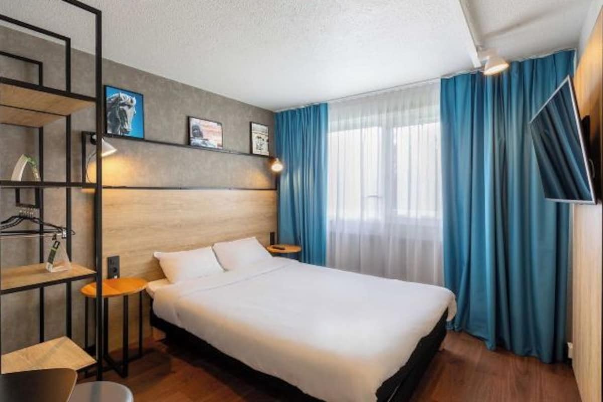 A hotel room with a bed, a desk, a chair, a shelf with items, and a window with curtains.