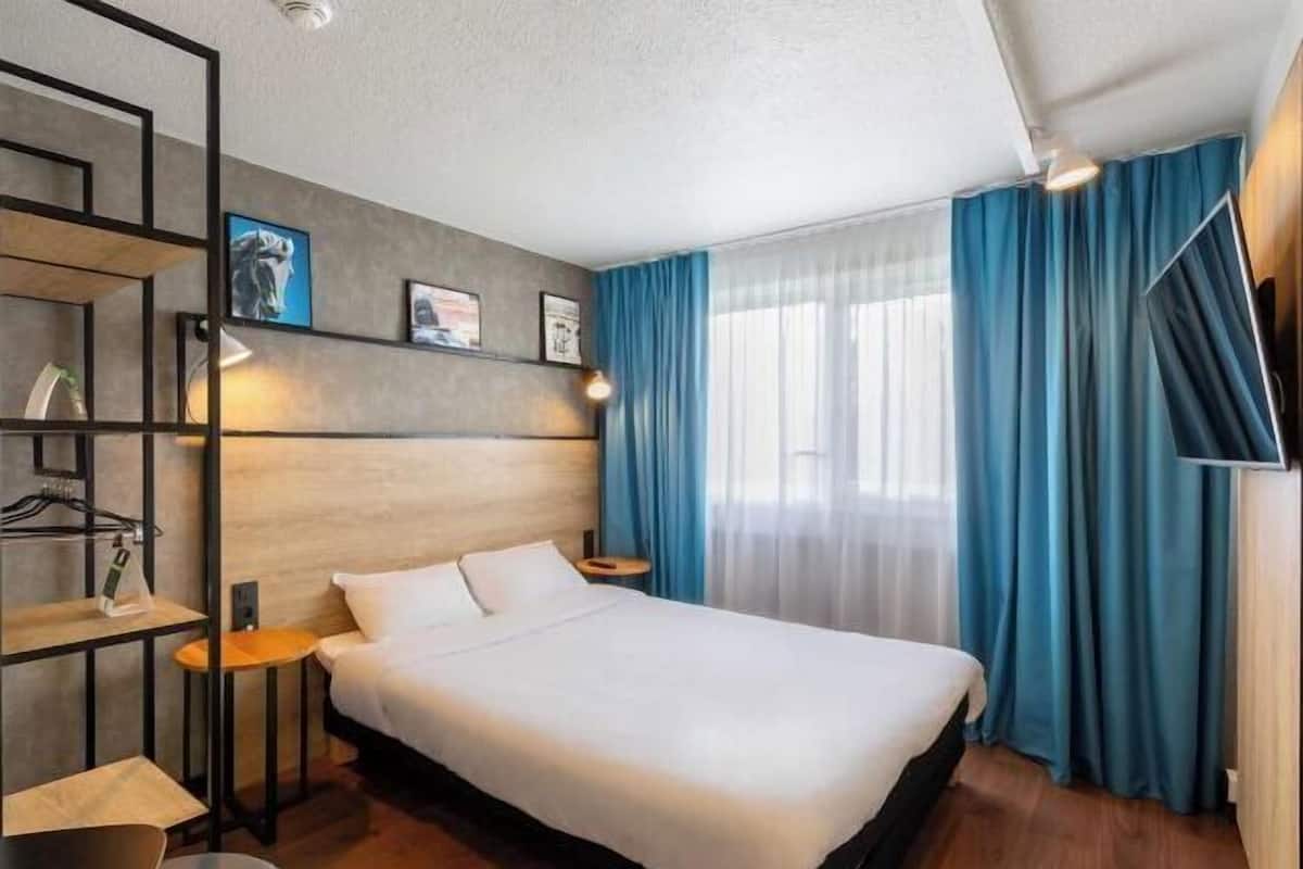 A hotel room with a bed, a desk, a chair, a shelf with items, and a window with curtains.