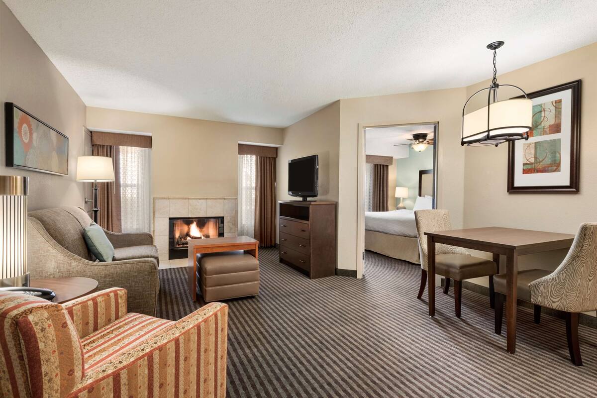 A hotel room with a sofa, armchair, ottoman, a small table, a TV, a fireplace, a desk with chairs, and a mirror.