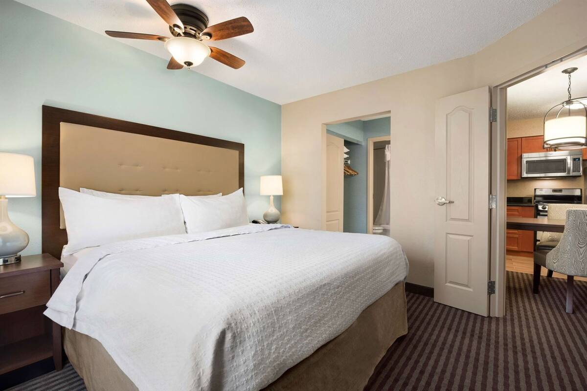 A hotel room with a bed, a ceiling fan, a nightstand, a lamp, a door leading to a bathroom, and a kitchenette.