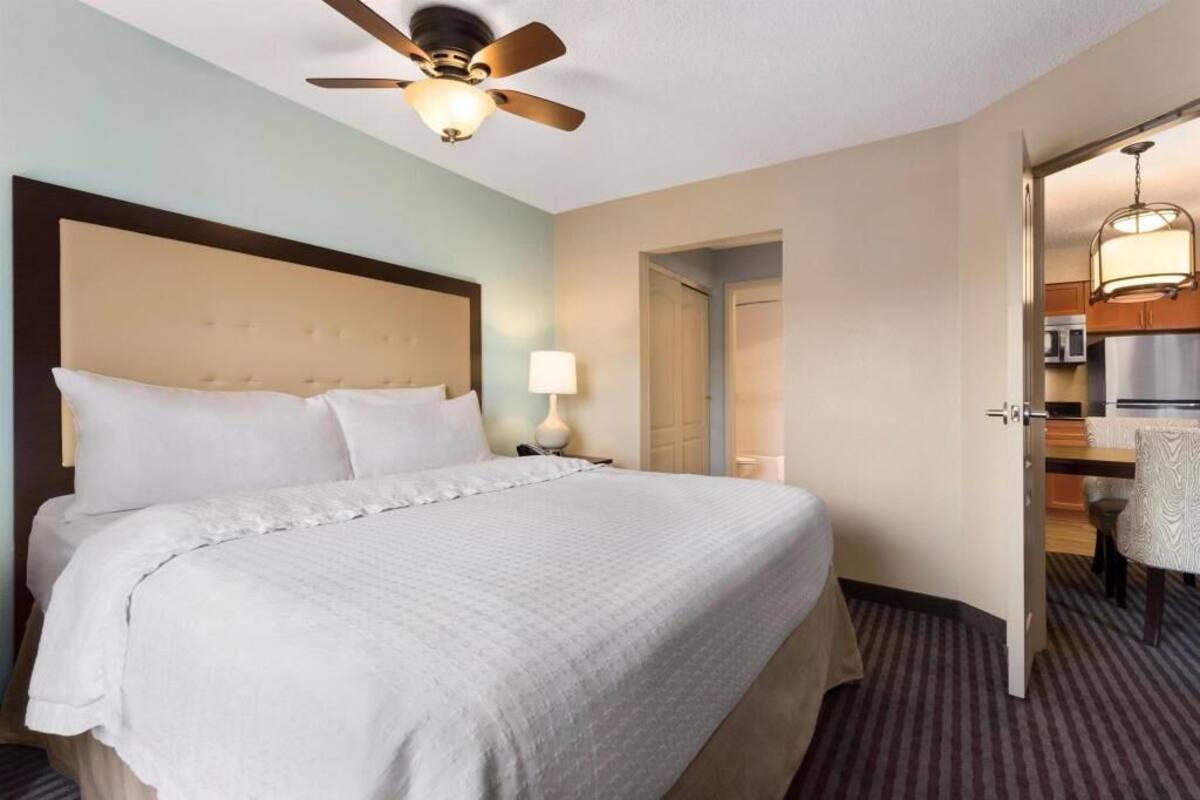 A hotel room with a bed, a ceiling fan, a nightstand, a lamp, and an open door leading to a kitchenette.