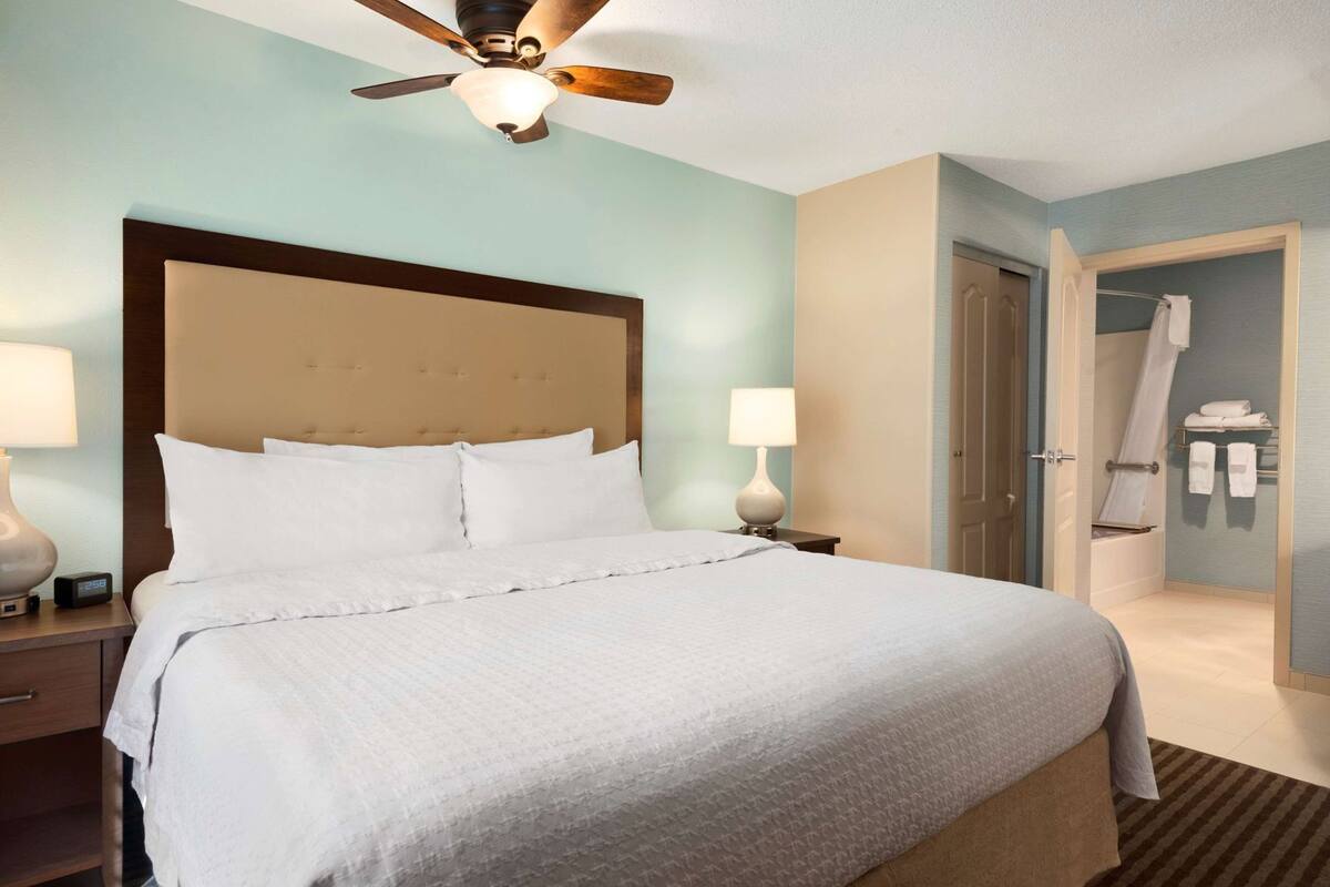 A hotel room with a large bed, a ceiling fan, a nightstand with a lamp, and a bathroom visible through an open door.