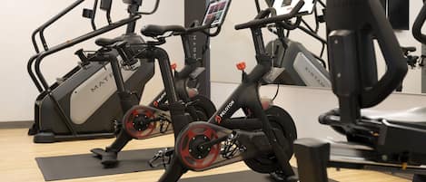 Three stationary bikes with digital screens, positioned on black mats in a room with wooden flooring and a large mirror.
