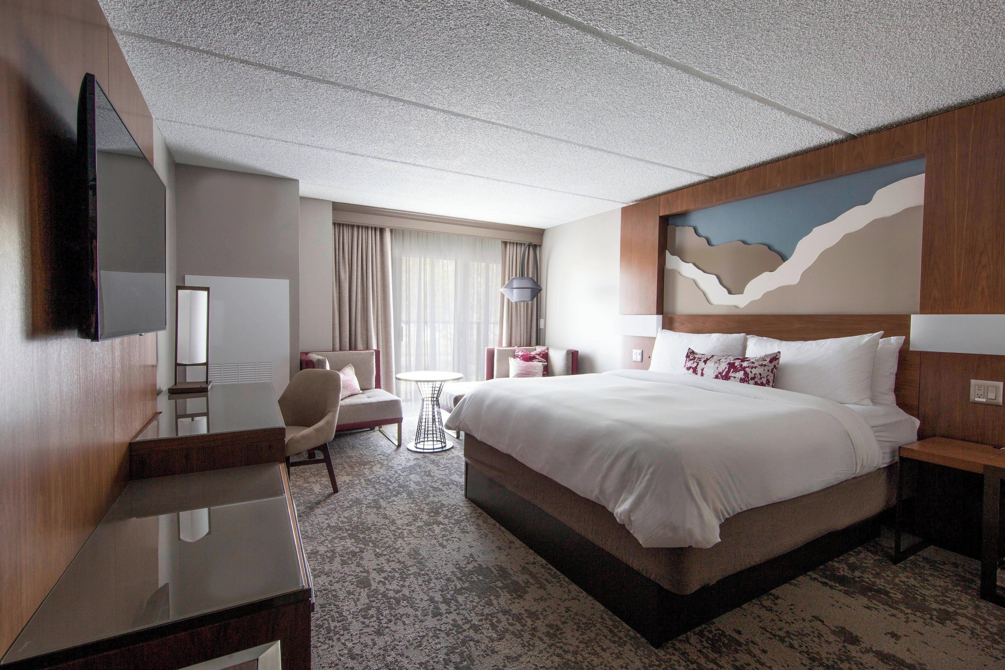 A hotel room with a large bed, a desk, a chair, a TV, and a window with curtains.