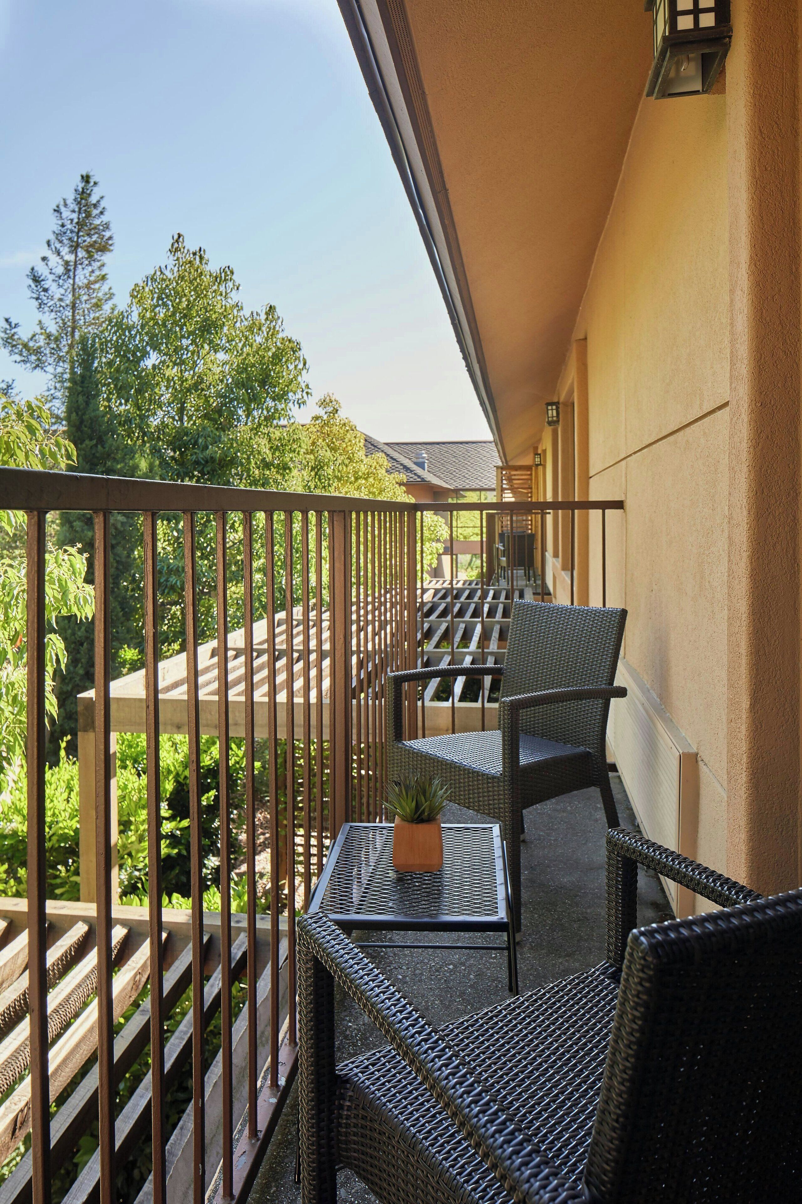 A balcony with wicker furniture and a small table, overlooking a garden and a building.