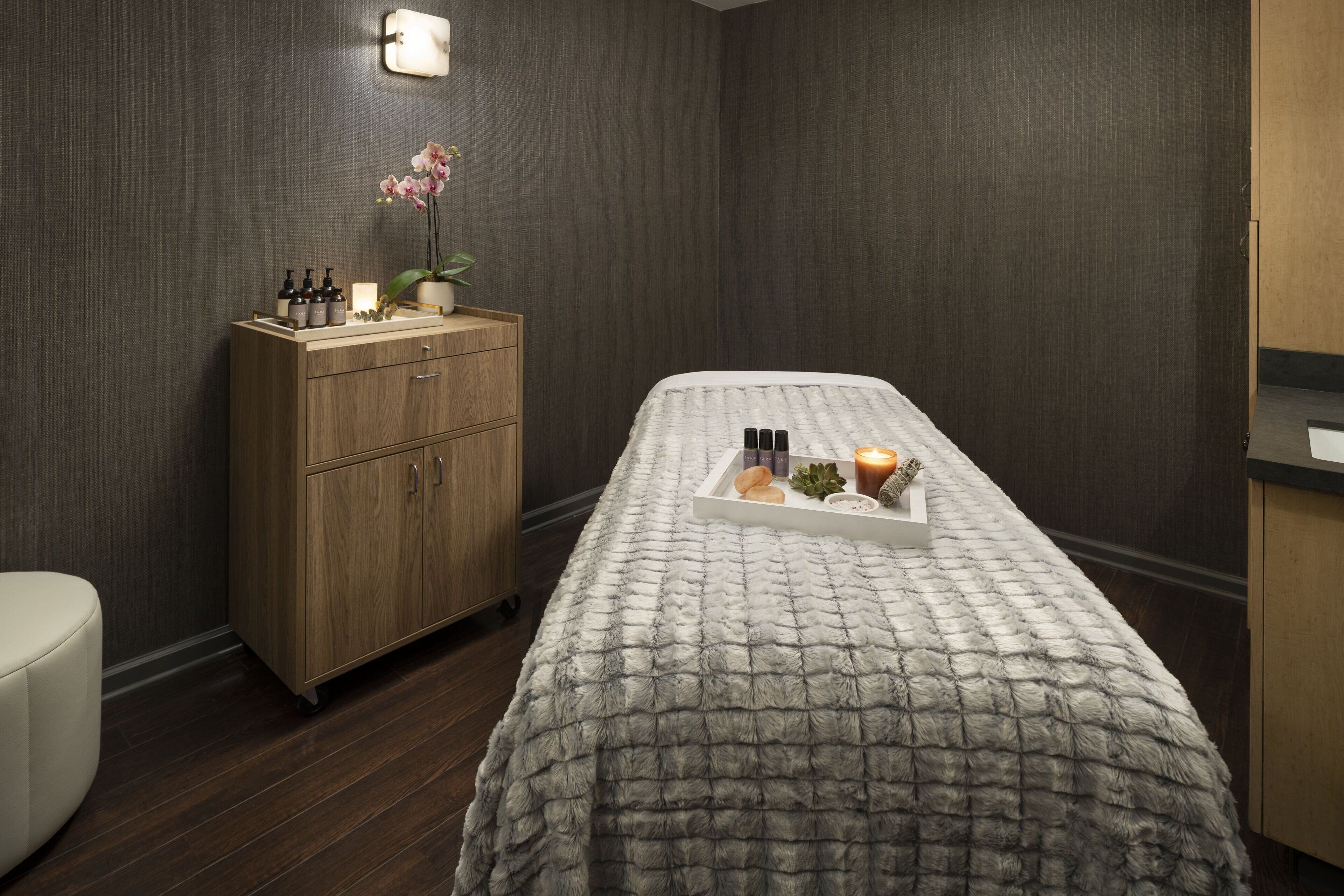 Un cuarto de spa con una camilla de masaje, un mueble de madera con botellas y una bandeja con velas y frutas.