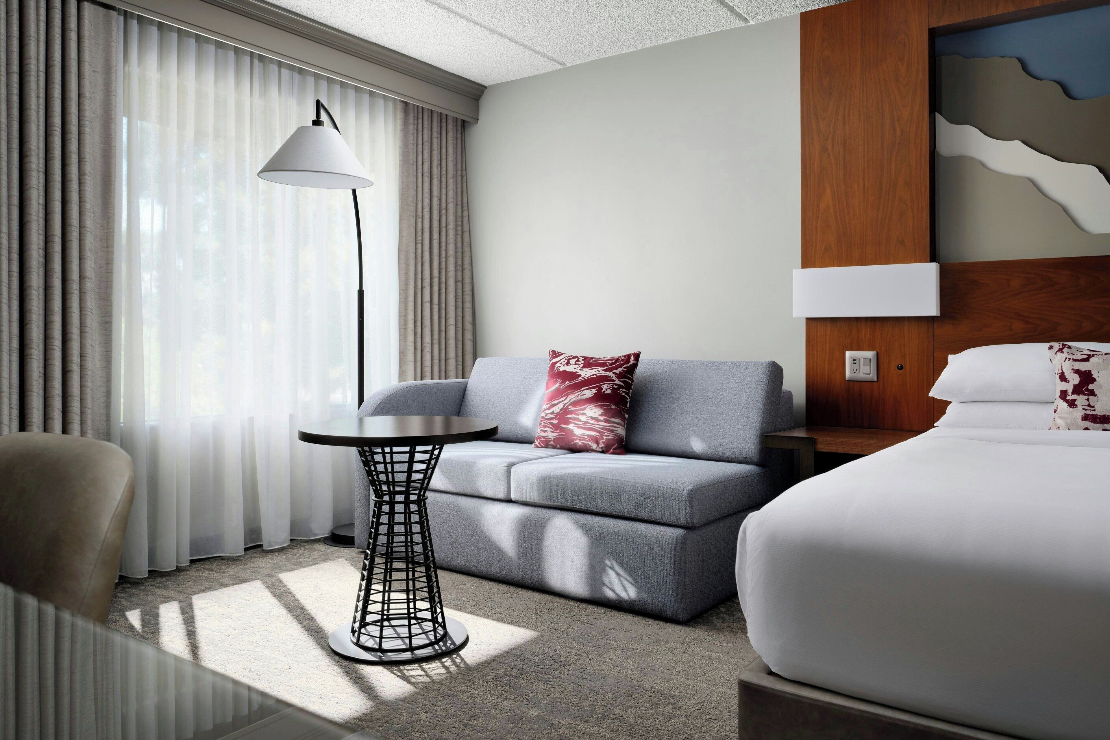 A hotel room with a grey sofa, a small round table, a lamp, a bed, and a wall with a wooden panel and a white abstract design.