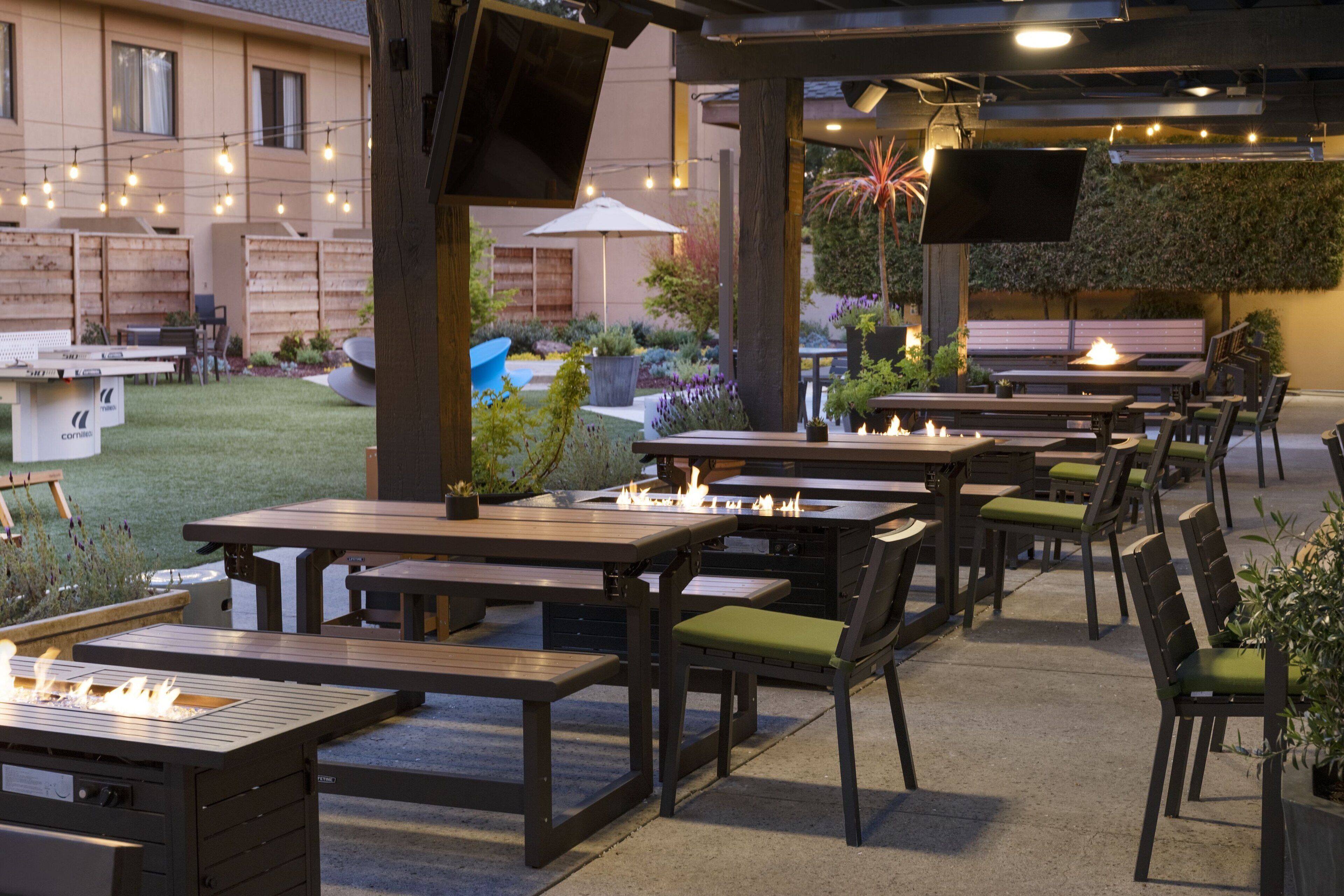 Un área de patio al aire libre con varias mesas y sillas de madera, un pozo de fuego y luces colgantes.