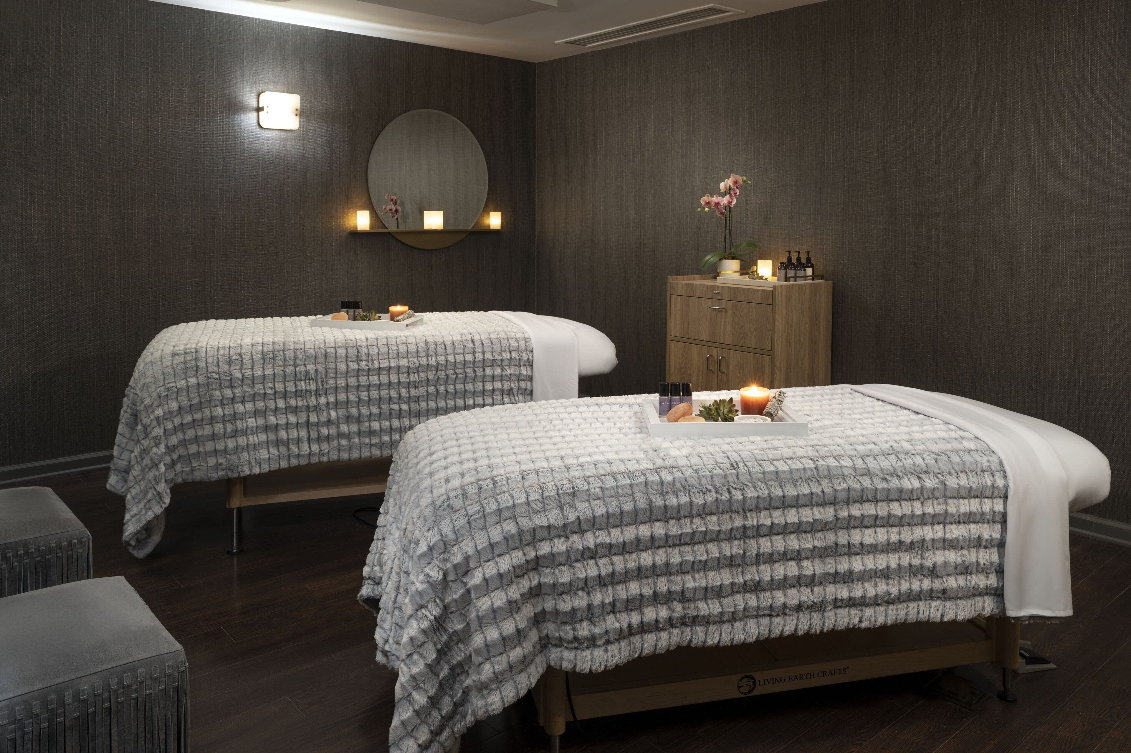 Twee bedden met grijs beddengoed met een patroon, een houten kastje met toiletartikelen, en een spiegel met verlichting die aan de muur is bevestigd.