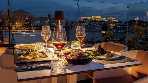 A table set for dinner with wine glasses, plates of food, and a view of a cityscape at dusk. - Brown Acropol, a Member of Brown Hotels (Athens)