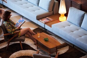 A person seated on a chair reading a magazine, with a sofa and a table with a lamp in the background. - Brown Acropol, a Member of Brown Hotels (Athens)