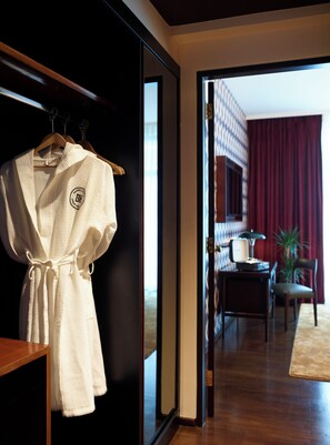 A hotel room with a wooden floor, a robe hanging on a wooden hanger, a desk with a lamp, and a chair. - Brown Acropol, a Member of Brown Hotels (Athens)