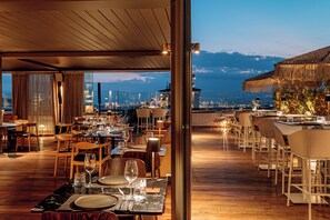 Un restaurante con mesas dispuestas para comer, vista del paisaje urbano y una montaña al fondo.