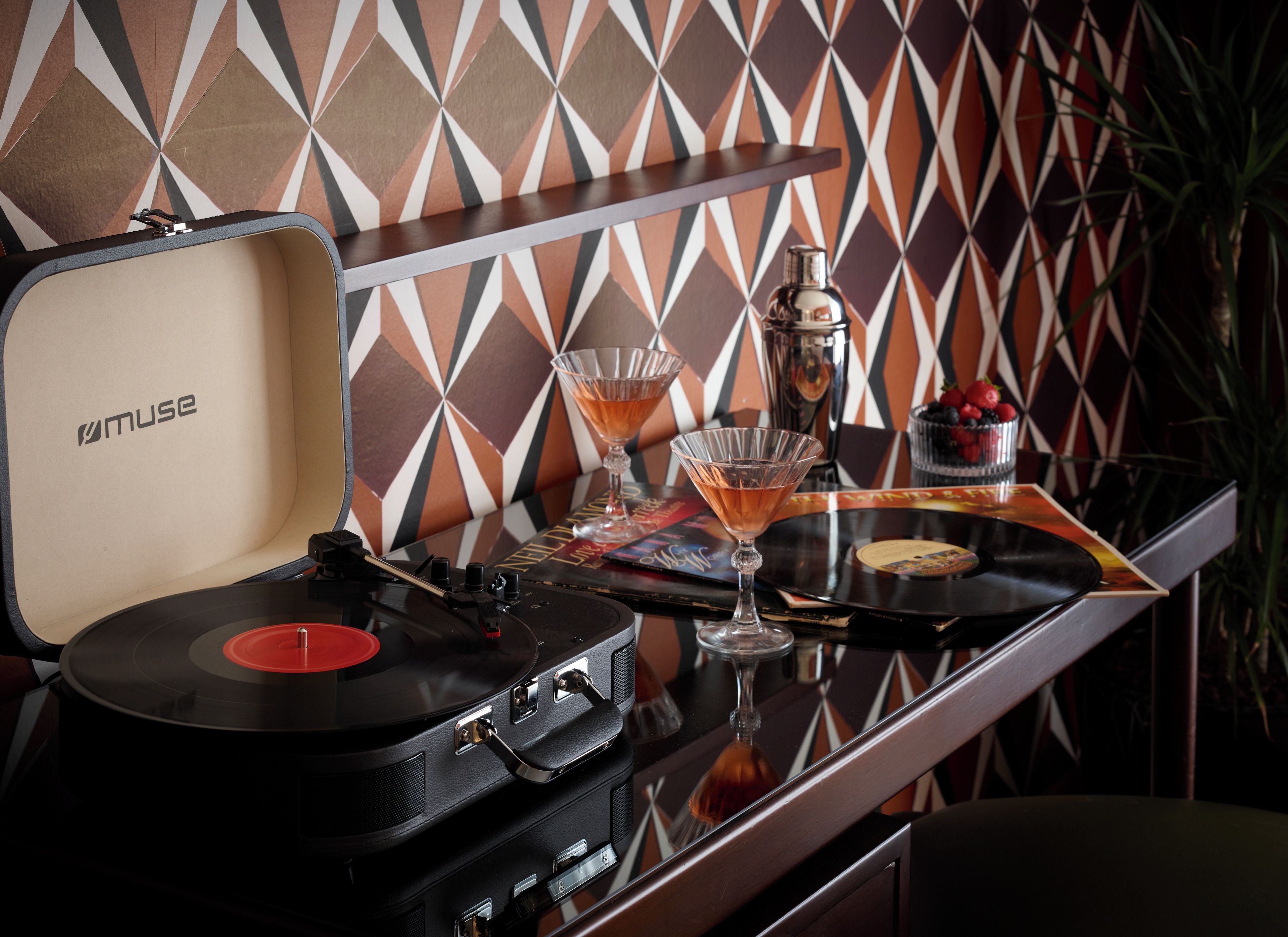 A turntable with a record, a martini glass, and a bottle on a table with a patterned wall in the background.