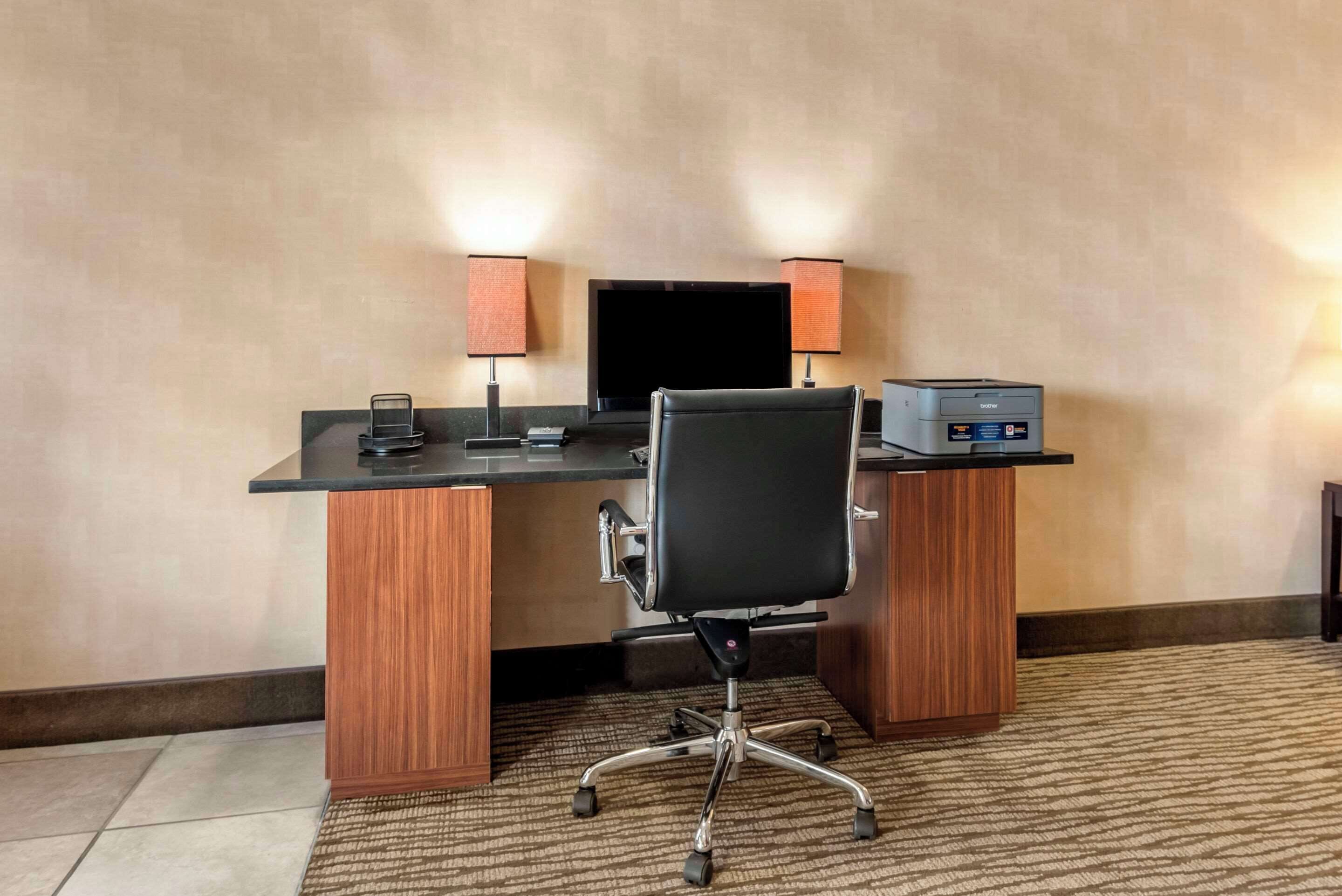 Habitación de hotel con un escritorio de madera, una silla de oficina negra, un televisor de pantalla plana, una impresora y dos lámparas.