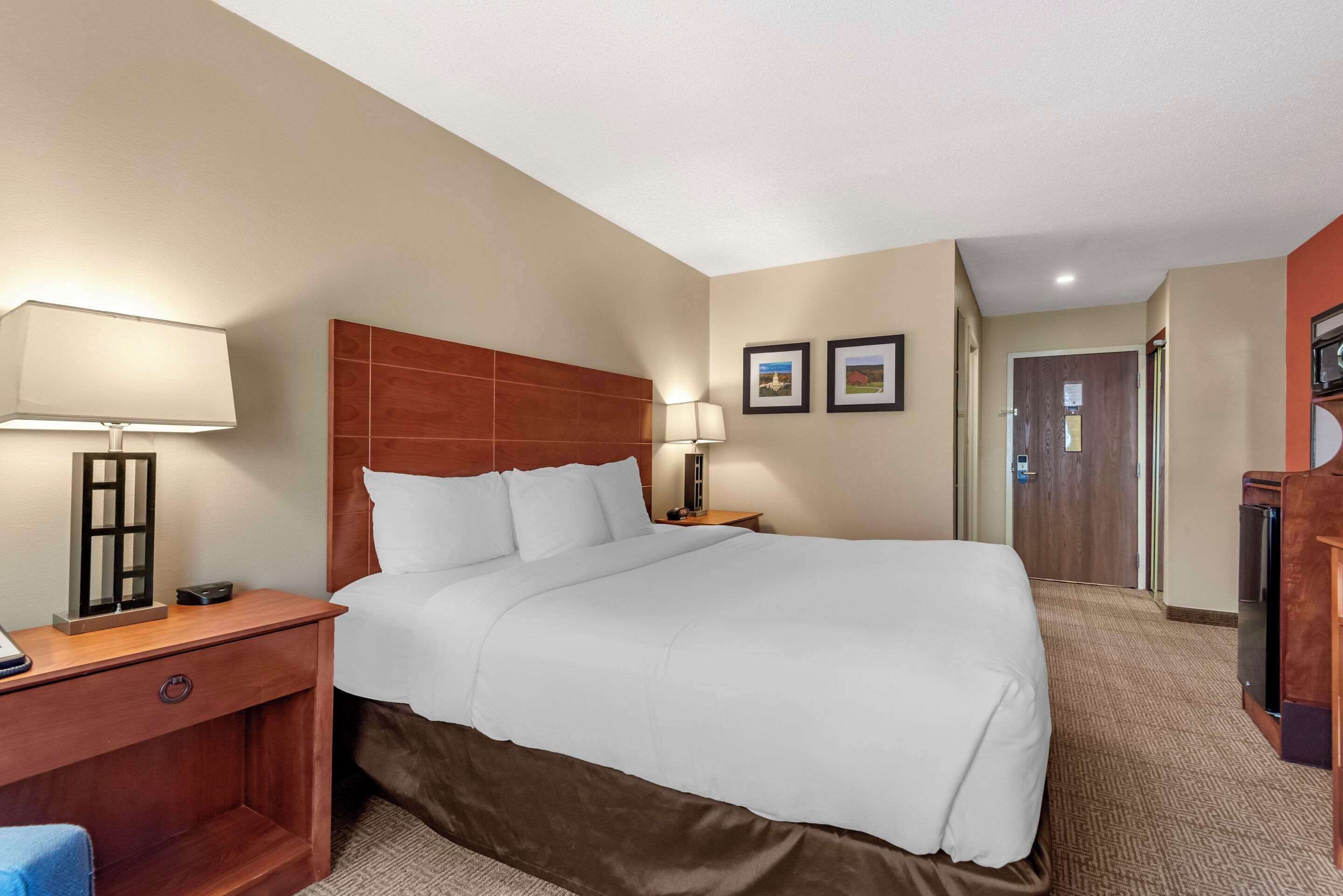 A hotel room with a large bed, a nightstand with a lamp, a wooden headboard, a wall-mounted TV, and a door leading to another room.