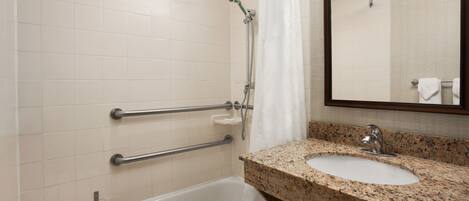 A bathroom with a white bathtub, a granite countertop sink, a mirror, and a shower with a handheld showerhead.