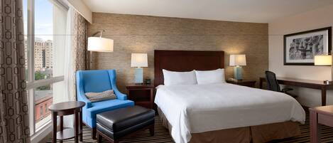 A hotel room with a large bed, a blue armchair, a small table, and a chair. There are two lamps on the nightstands and a window with curtains.