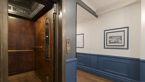Un ascensor con puerta de madera y panel de control, pared revestida de madera azul y un cuadro enmarcado.