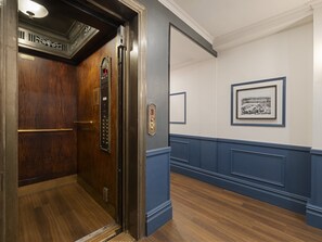 An elevator with a wooden door and a control panel, a blue wainscotted wall, and a framed picture on the wall. - voco The Franklin New York by IHG (New York)
