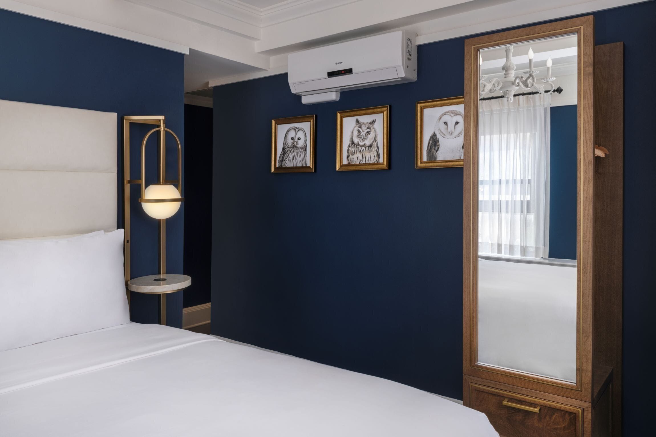 A hotel room with a bed, a round table with a lamp, a large mirror, and three framed owl pictures on the wall.