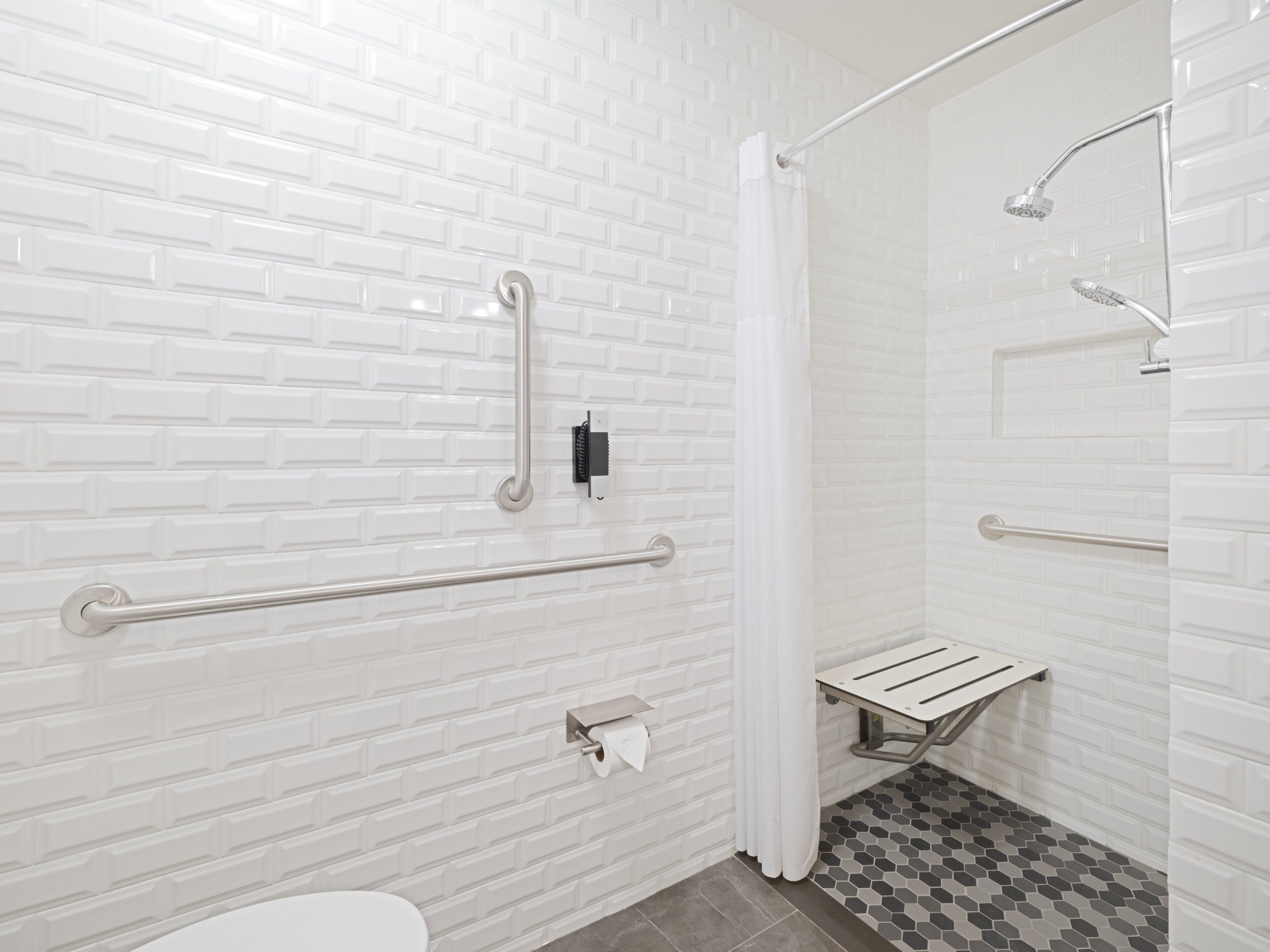 A bathroom with white brick walls, a shower with a handheld showerhead, a toilet, and a grab bar.